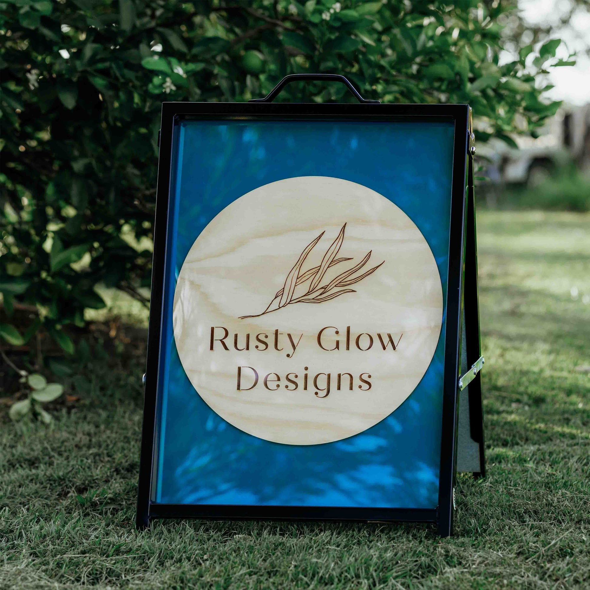 A sign with 'Rusty Glow Designs' logo on a grassy background. This is a sign that is an example of the type of A-frame signs that can be custom made.