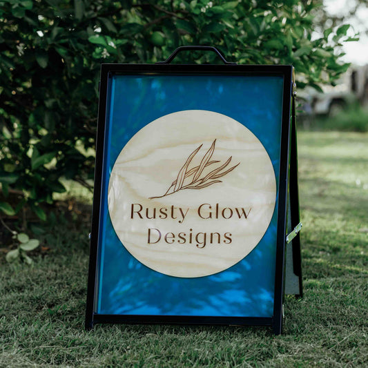 A sign with 'Rusty Glow Designs' logo on a grassy background. This is a sign that is an example of the type of A-frame signs that can be custom made.